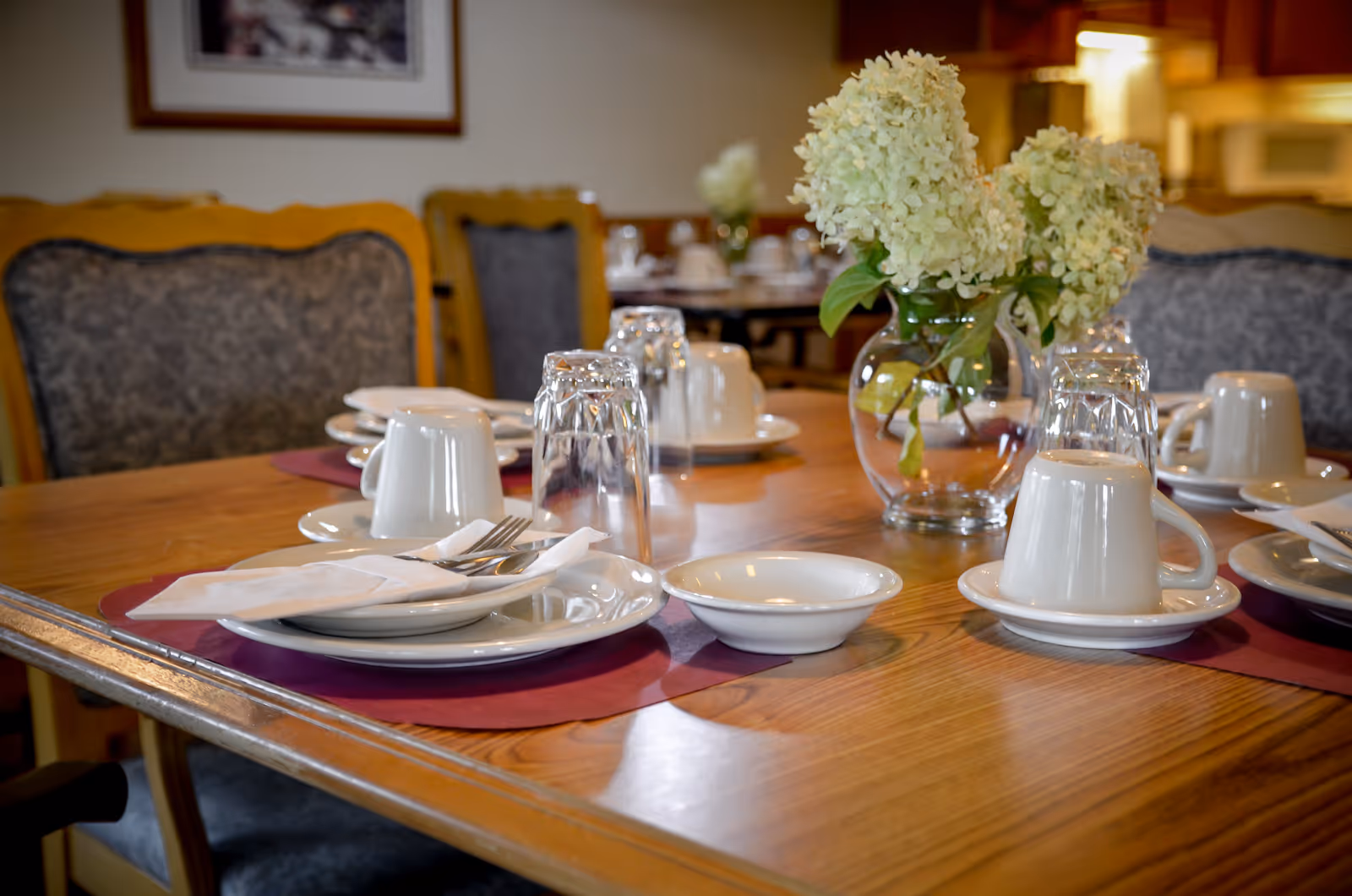 A dining table set with plates, cups, glasses, utensils and a vase of white flowers in a communal dining room.
