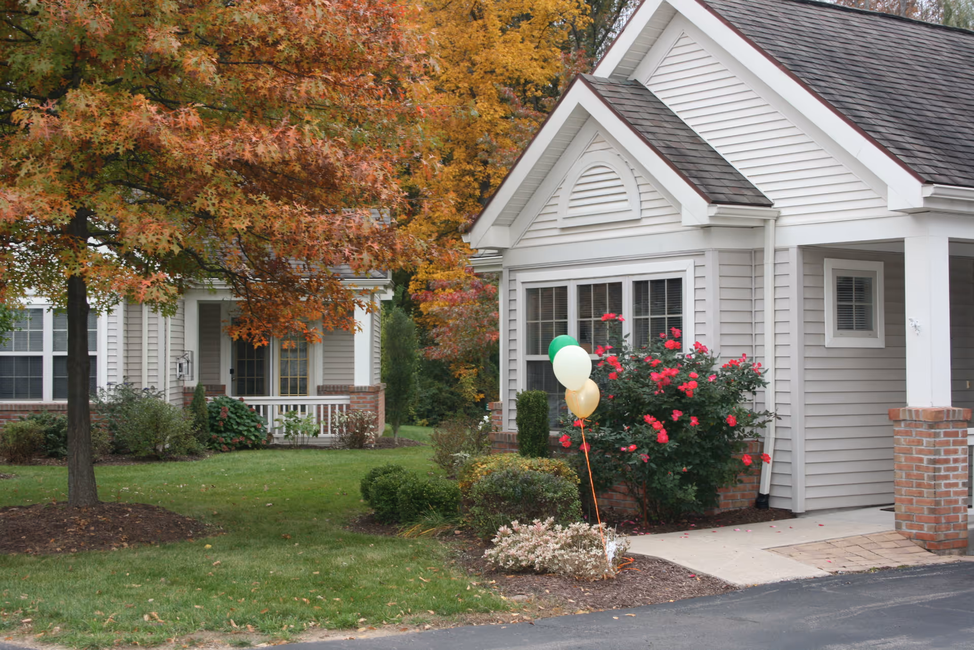 Exterior view of a residential building with beige siding and white trim, surrounded by green grass, bushes, and a tree with autumn-colored leaves. There are three balloons (green, white, and gold) tied to a bush with red flowers near the building entrance.