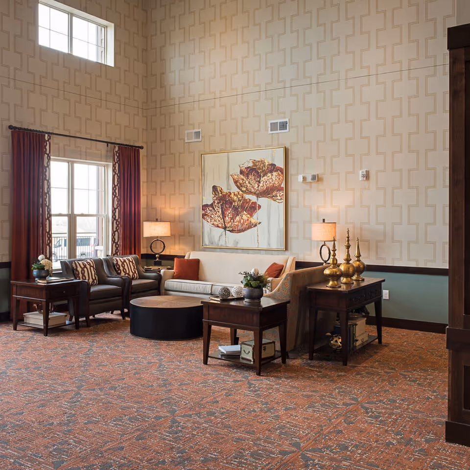 A cozy senior living facility lounge area with a beige sofa, two dark leather armchairs, a round coffee table, and two wooden side tables. The walls have a patterned wallpaper, and there is a large painting of flowers above the sofa. Two table lamps with beige shades provide warm lighting. The room has large windows with red curtains and a high ceiling.
