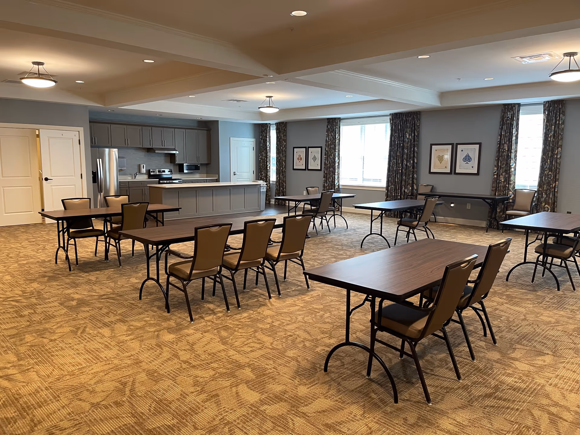 A spacious room with multiple rectangular tables and chairs arranged in rows on a patterned carpet. The room features a kitchen area with gray cabinets, a refrigerator, stove, and microwave. Large windows with floral curtains allow natural light to enter. The walls are decorated with framed artwork depicting playing card suits.