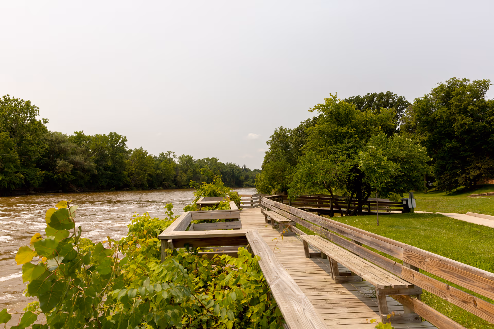 A wooden boardwalk with benches along a riverbank surrounded by green trees and bushes under a cloudy sky.
