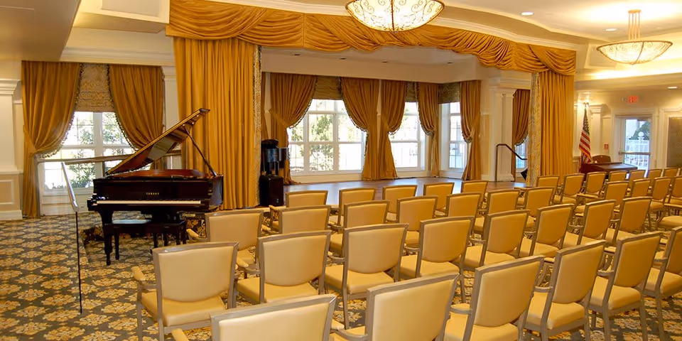 A well-lit event room with rows of beige chairs facing a grand piano on the left side. The room features large windows with golden curtains, patterned carpet, and elegant ceiling lights. An American flag is visible near the exit door on the right.