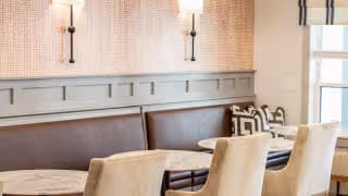 A cozy dining area with a built-in brown leather bench along the wall, beige upholstered chairs, and round marble-top tables. The wall behind the bench features decorative paneling and two wall-mounted light fixtures with white shades. A window with white blinds allows natural light into the space.