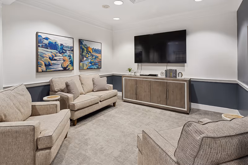 A cozy living room with beige upholstered sofas and armchairs arranged around a wooden cabinet with a large flat-screen TV mounted on the wall above it. The walls are painted white with a dark gray wainscoting, and two colorful landscape paintings hang on one wall. The room is carpeted and well-lit with recessed ceiling lights.