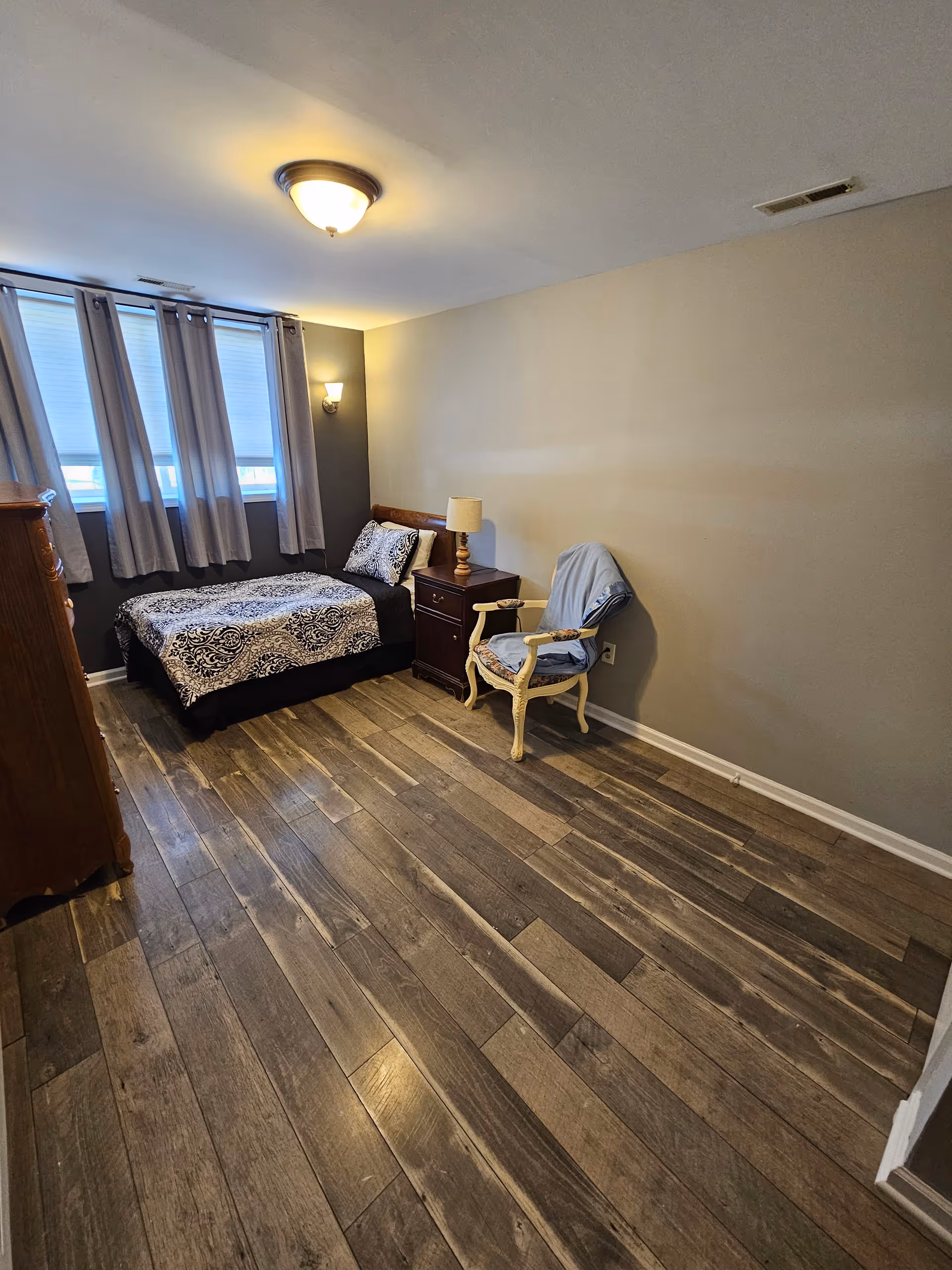 A small bedroom with a single bed covered in a black and white patterned bedspread, a wooden nightstand with a lamp, a vintage-style chair with a blue cover, wooden flooring, and three windows with gray curtains.