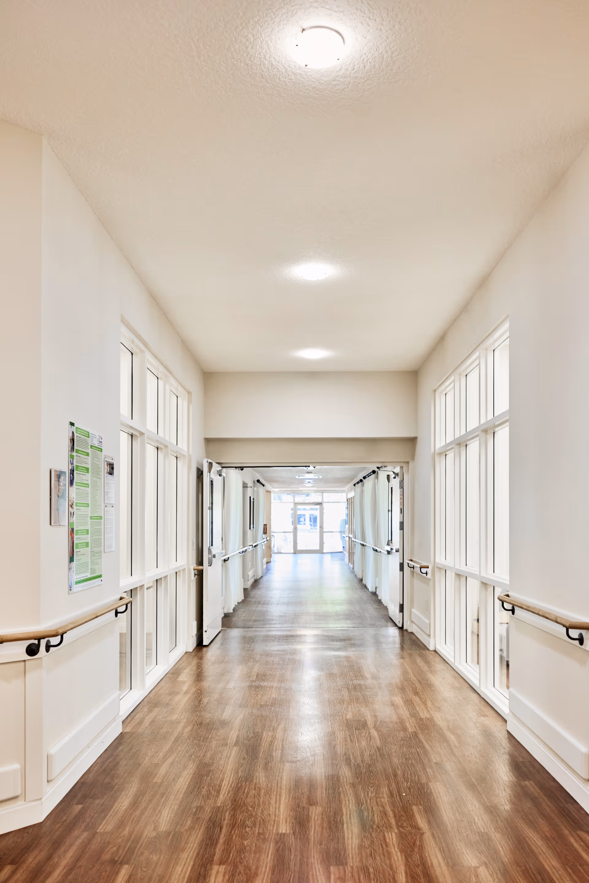A bright, clean hallway in a senior living facility with wooden floors, white walls, handrails on both sides, and large windows along the corridor. The hallway leads to a glass door at the far end, allowing natural light to enter.