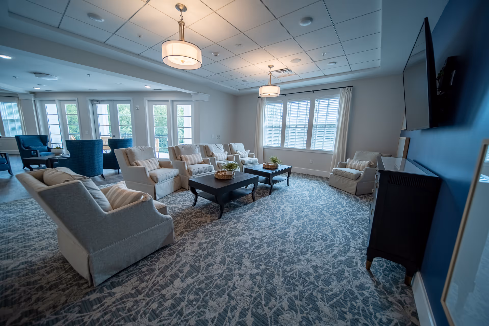 A spacious and well-lit living room with multiple beige armchairs arranged around two dark wooden coffee tables. The room features large windows with white blinds and curtains, a patterned carpet, two ceiling light fixtures, and a wall-mounted flat-screen TV on a blue accent wall. Additional seating with blue chairs is visible near glass doors leading outside.