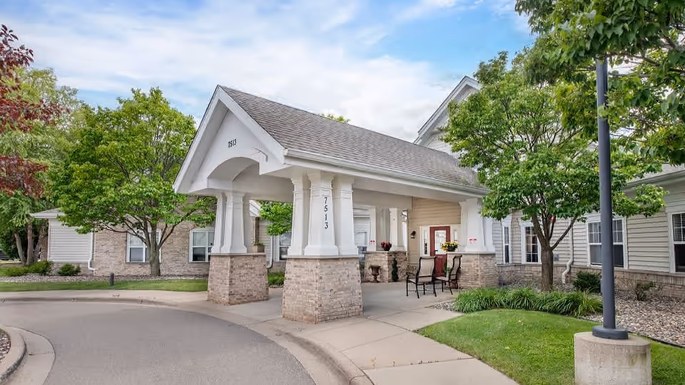 Covered entrance/porte-cochere of a senior living building with brick columns, seating, and surrounding trees and landscaping.