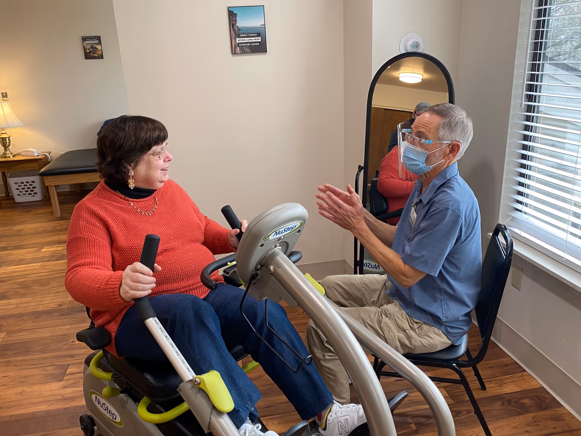 A woman in a red sweater is using a NuStep exercise machine while a man wearing a blue shirt, face mask, and face shield sits on a chair in front of her, clapping his hands. They are in a room with wooden floors, a window with blinds, a mirror, and a small table with a lamp in the background.