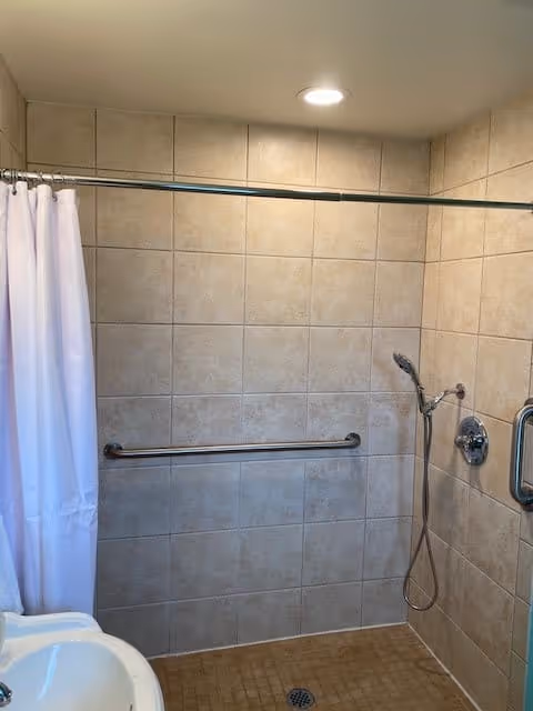 A tiled bathroom shower area with beige tiles on the walls and floor, a white shower curtain on a curved rod, a handheld showerhead attached to the wall, a grab bar for support, and a ceiling light above.