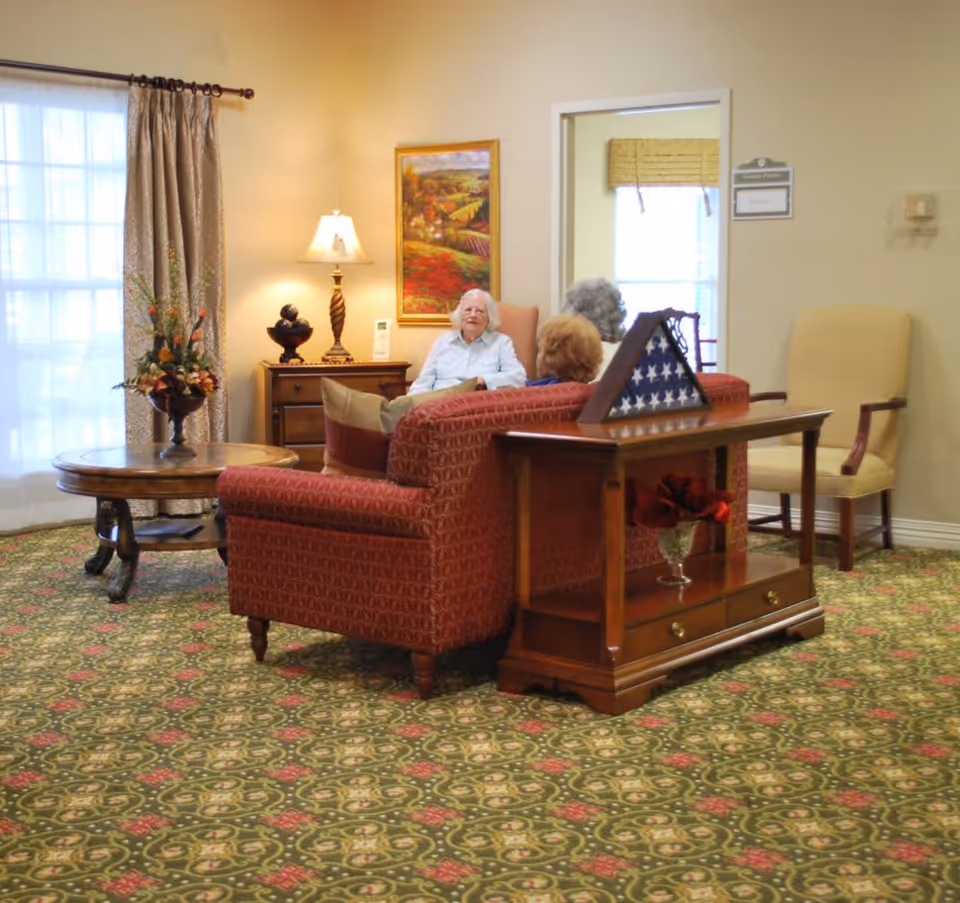 A cozy living room area in a senior living facility with three elderly women sitting and conversing on a red patterned sofa. The room features a patterned carpet, a wooden side table with a folded American flag display, a round coffee table with a floral arrangement, a lamp on a wooden cabinet, and a framed landscape painting on the wall. Light filters in through a window with curtains.