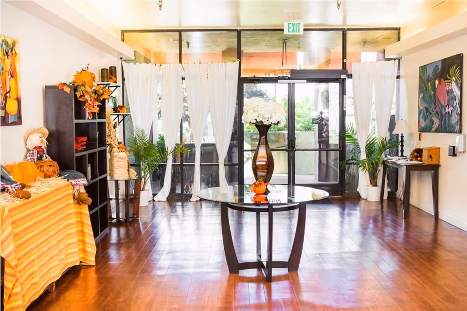 Bright senior living facility entrance area with glass double doors, sheer white curtains, a round glass table with a tall vase of white flowers, potted plants, seasonal decorations including pumpkins and a scarecrow, and colorful artwork on the walls.