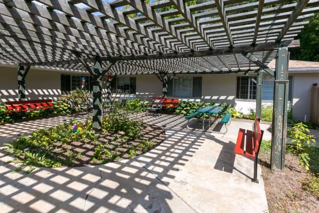 Shaded outdoor courtyard with a wooden pergola casting lattice shadows over benches, a picnic table, and planted garden beds beside a single-story building.