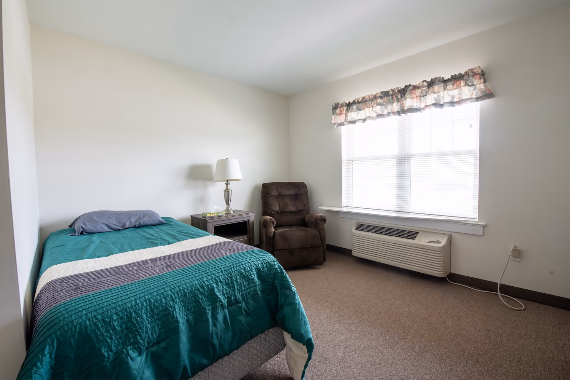 A simple bedroom with a single bed covered in a teal, white, and gray striped comforter, a gray pillow, a wooden nightstand with a lamp and a box of tissues, a brown recliner chair, a large window with blinds and a floral valance, and a wall-mounted air conditioning unit below the window.
