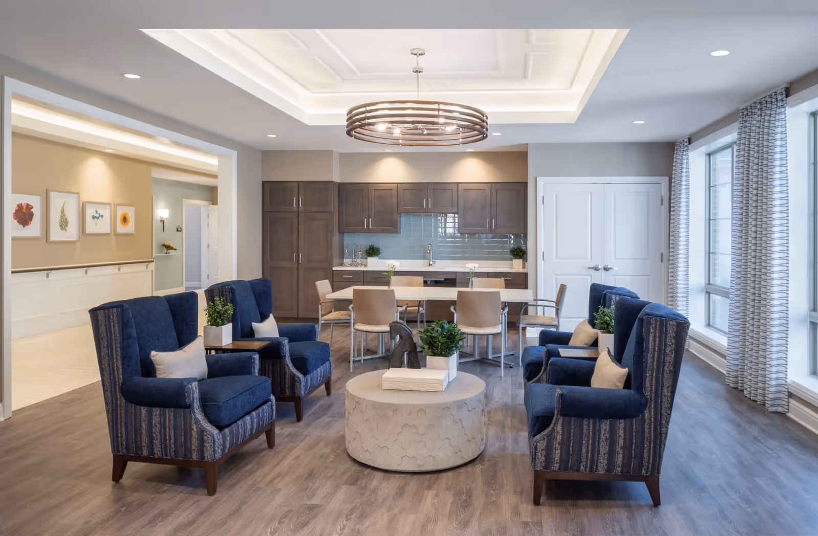 Bright common living area with blue armchairs arranged around a round coffee table facing a kitchen/dining area and large windows.