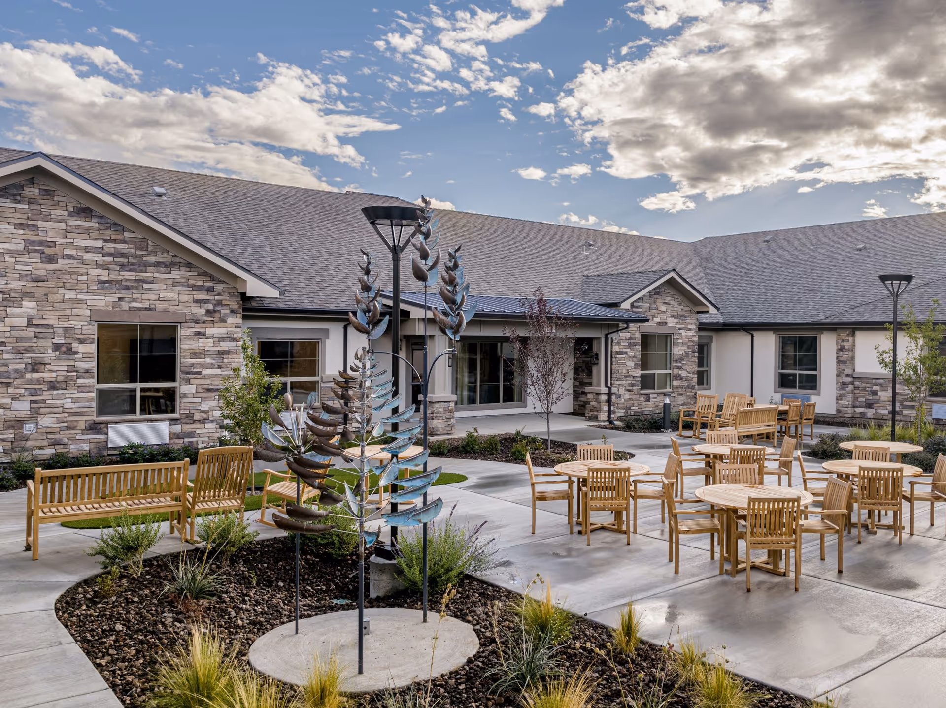 Outdoor courtyard area of Kiley Ranch Senior Living featuring wooden benches and round tables with chairs on a concrete patio, surrounded by landscaped plants and a decorative metal sculpture, with a stone and stucco building in the background under a partly cloudy sky.