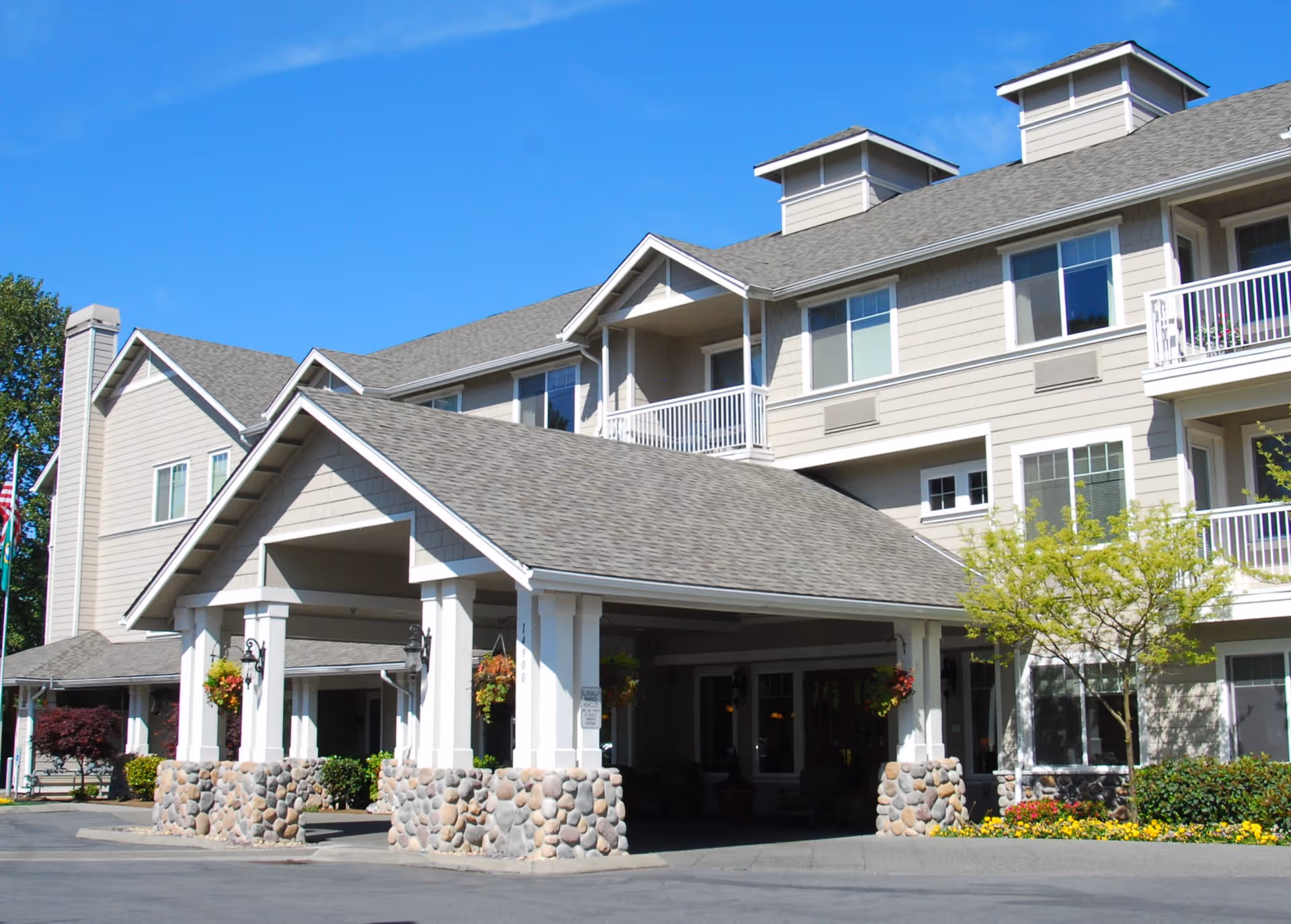 Exterior view of a multi-story senior living facility with a covered entrance supported by white columns with stone bases, surrounded by landscaping including trees and flowers under a clear blue sky.