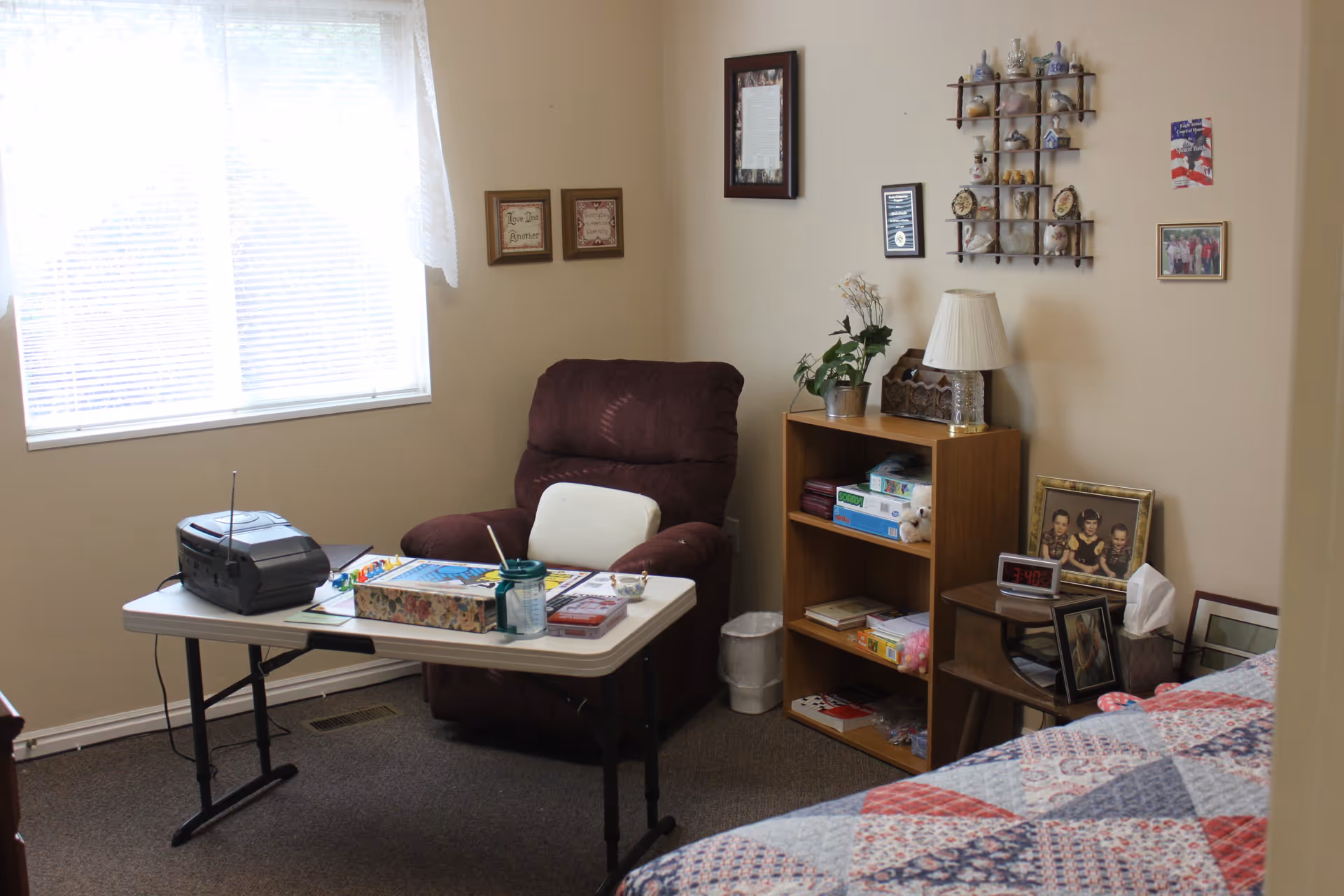 A cozy room with a large window covered by sheer curtains, a brown recliner chair, a folding table with a radio, board games, and a cup. There is a wooden bookshelf filled with board games and decorative items, a small side table with framed photos, a digital clock showing 3:40, a tissue box, and a bed with a colorful quilted bedspread. The walls are decorated with framed pictures and a small shelf holding various knick-knacks.
