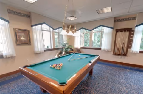 A well-lit game room featuring a green felt pool table with billiard balls arranged in a triangle and two pool cues resting on the table. The room has large windows with white curtains, a hanging light fixture above the table, a rack with additional pool cues on the wall, and a framed picture on another wall. The floor is carpeted with a blue patterned carpet.
