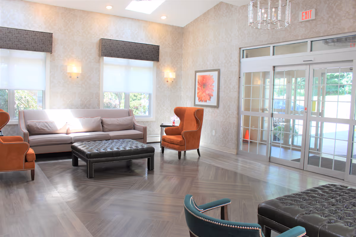 A bright and spacious seating area with a beige sofa, two orange armchairs, a teal chair, and a large black tufted ottoman. The room has patterned wallpaper, two windows with white blinds and dark valances, a framed flower artwork on the wall, and glass double doors leading outside. The floor is wood with a herringbone pattern and there is a chandelier hanging from the ceiling.