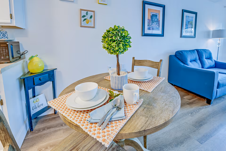 A small round wooden dining table set for two with white bowls, plates, mugs, and silverware on orange and white placemats. A small potted green plant is in the center of the table. In the background, there is a blue couch, framed pictures on the wall, a floor lamp, and a small blue side table with a yellow decorative item and a sign that says 'kitchen'.