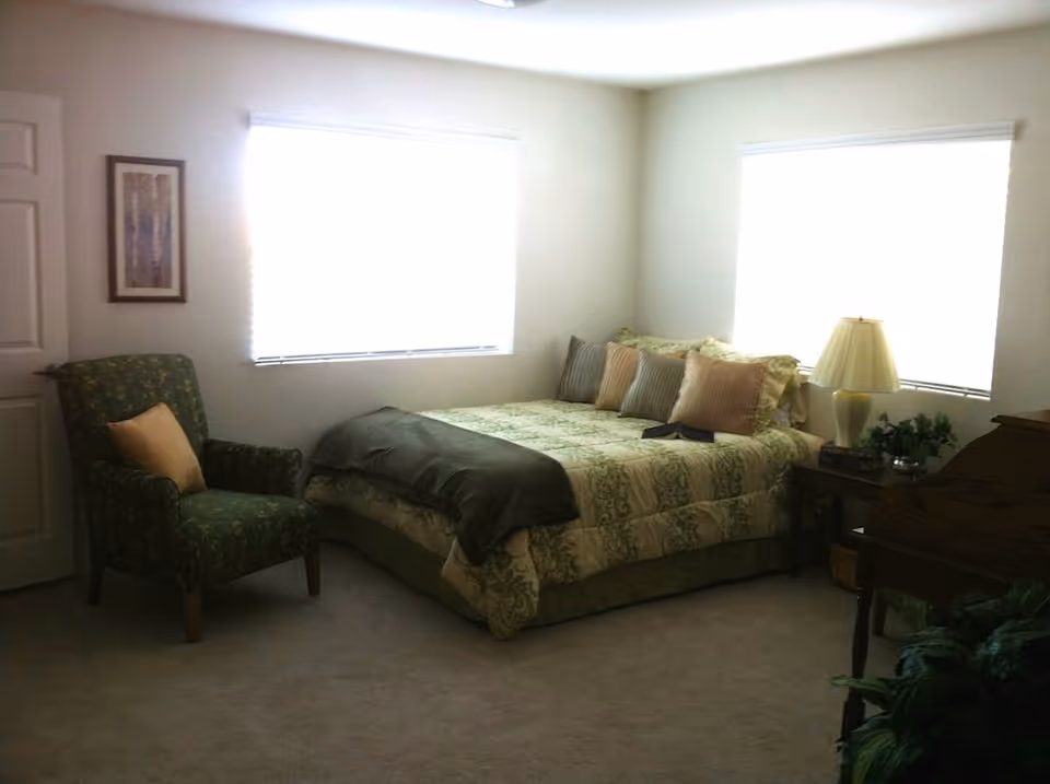 A tidy bedroom with a bed, chair, nightstand, lamp, piano, and two windows.