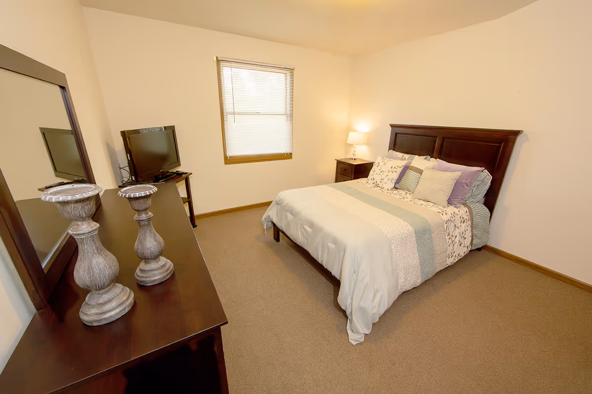 A simple furnished bedroom with a bed, nightstand and lamp, dresser with mirror and TV, and a window.