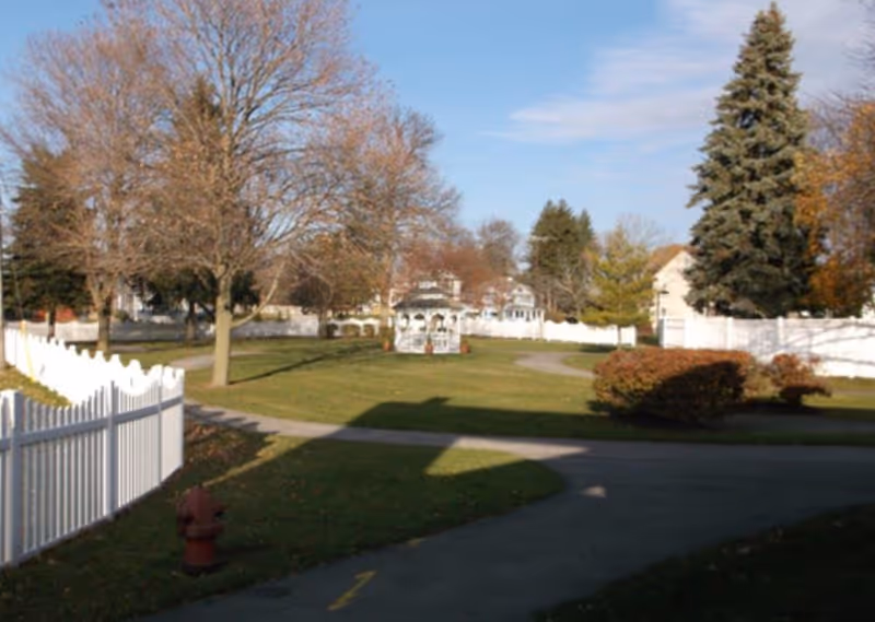 Outdoor area with a paved walking path, green grass, leafless trees, a white gazebo in the distance, white picket fences, and some bushes under a partly cloudy sky.