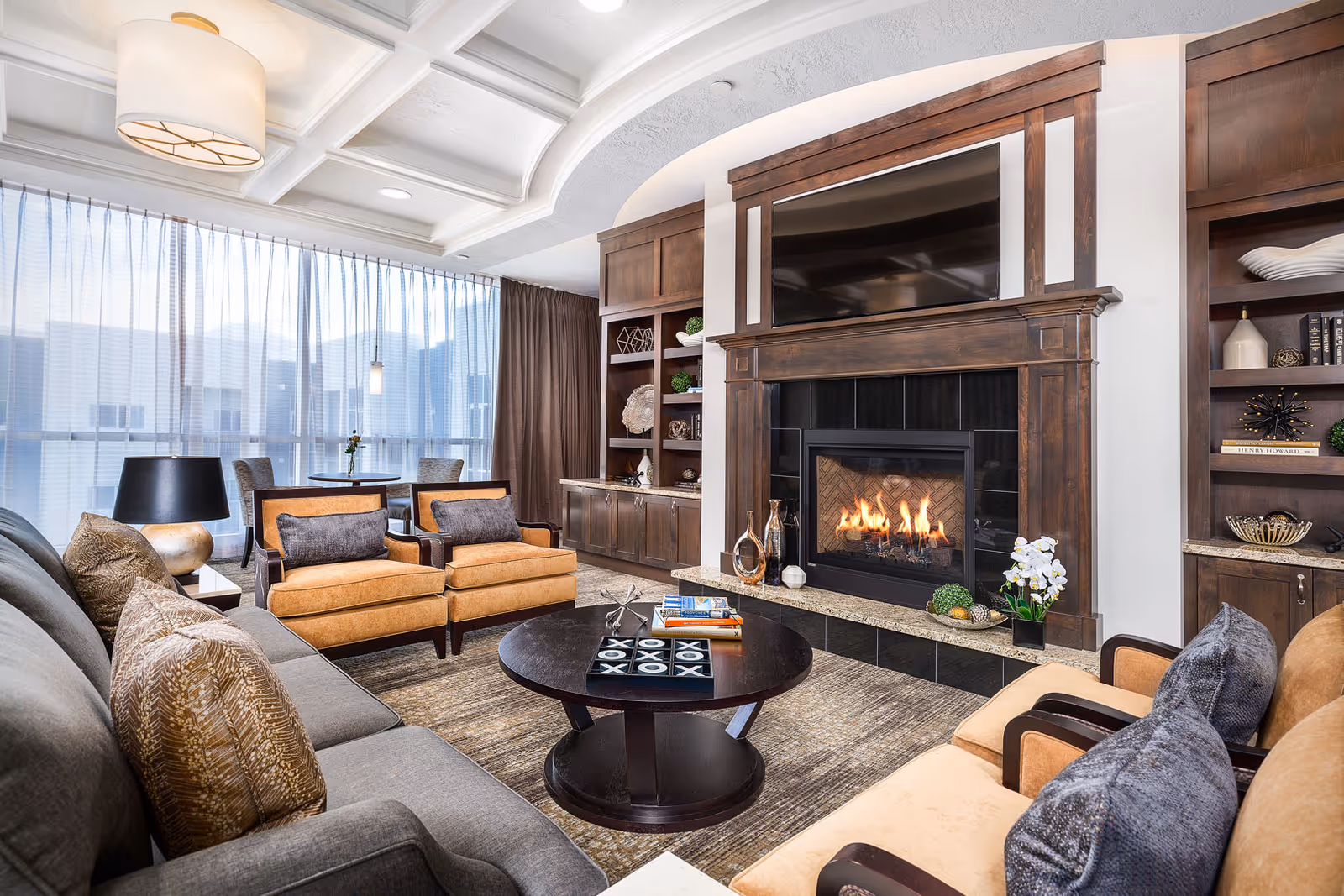 A cozy living room with a lit fireplace surrounded by dark wood cabinetry and a mounted flat-screen TV above it. The room features a gray sofa with patterned pillows, two tan armchairs with dark cushions, a round dark wood coffee table with books and a tic-tac-toe game, and a small dining table with chairs near large windows covered by sheer curtains. The ceiling has a decorative coffered design with a large drum light fixture.