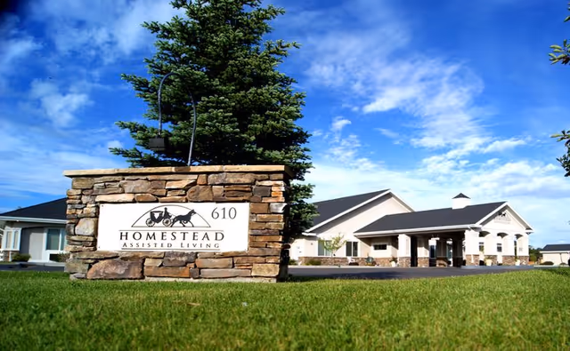 Stone entrance sign reading "Homestead Assisted Living 610" in front of a single-story assisted living building under a blue sky.