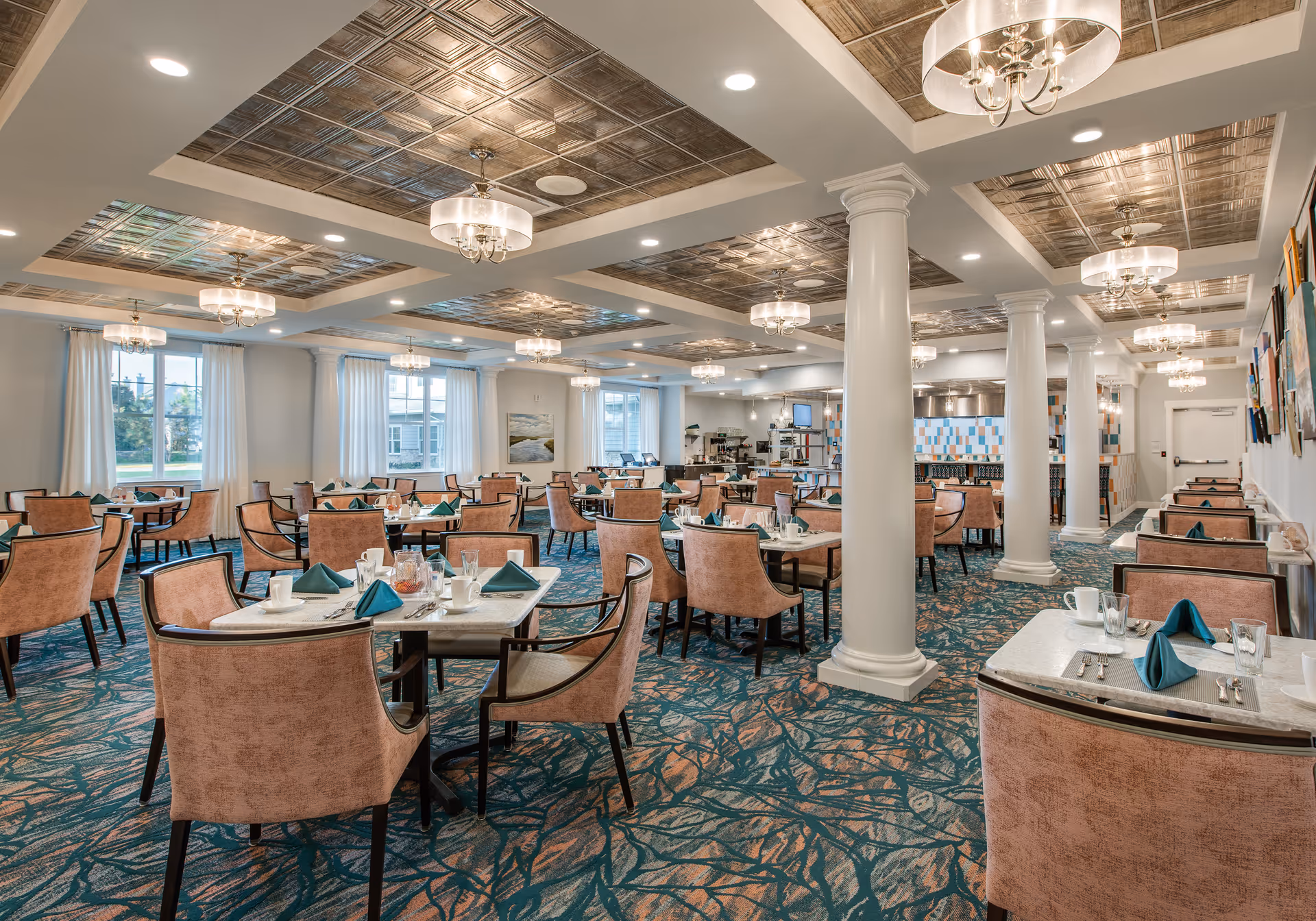 Spacious dining room with multiple tables set with white plates, cups, silverware, and teal napkins. The room features pink upholstered chairs, patterned carpet, white pillars, large windows with white curtains, and decorative ceiling panels with chandeliers.