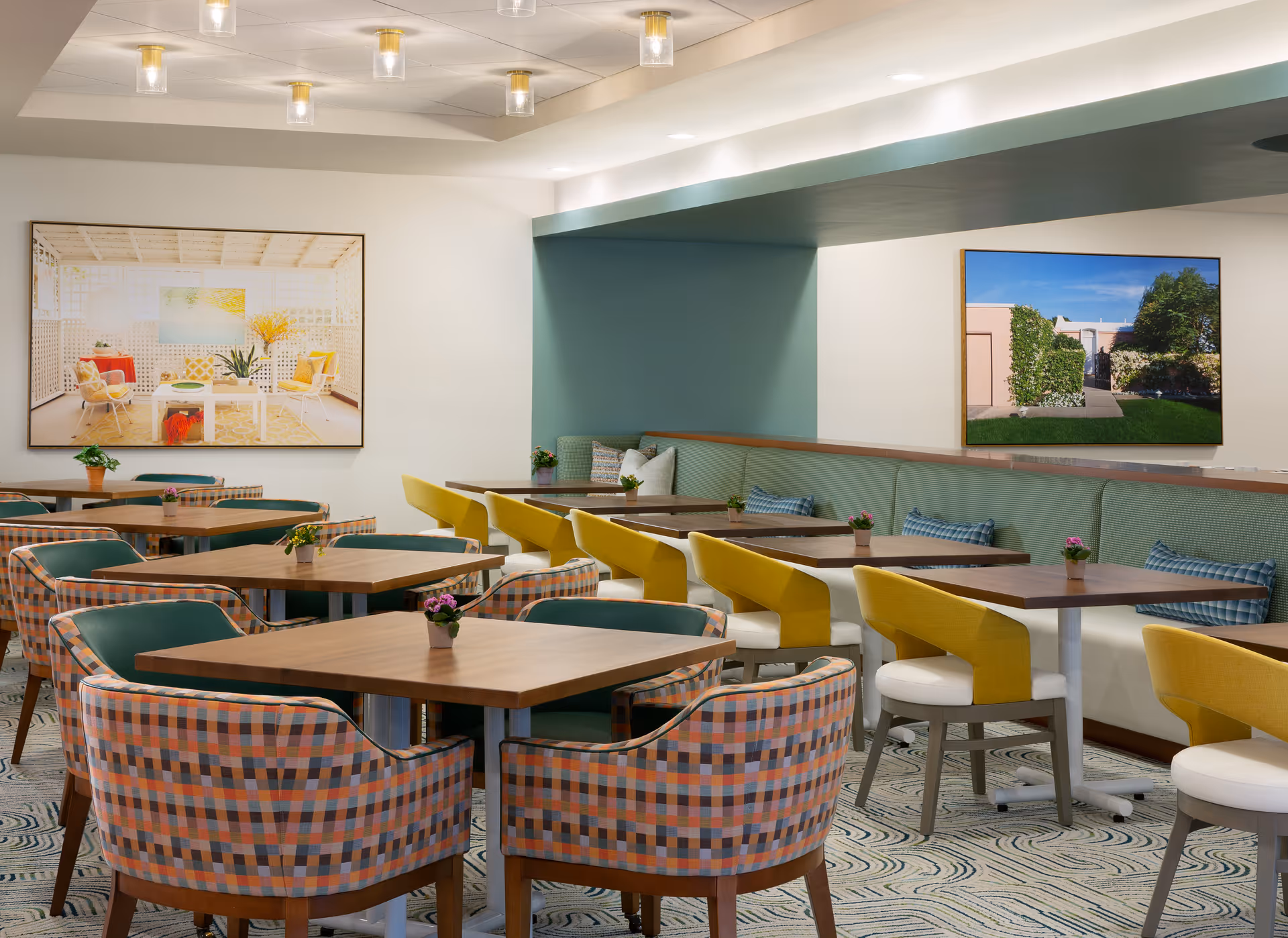 Bright, modern dining room with wooden tables, patterned armchairs, yellow and white chairs, a teal banquette, and framed artwork on the walls.