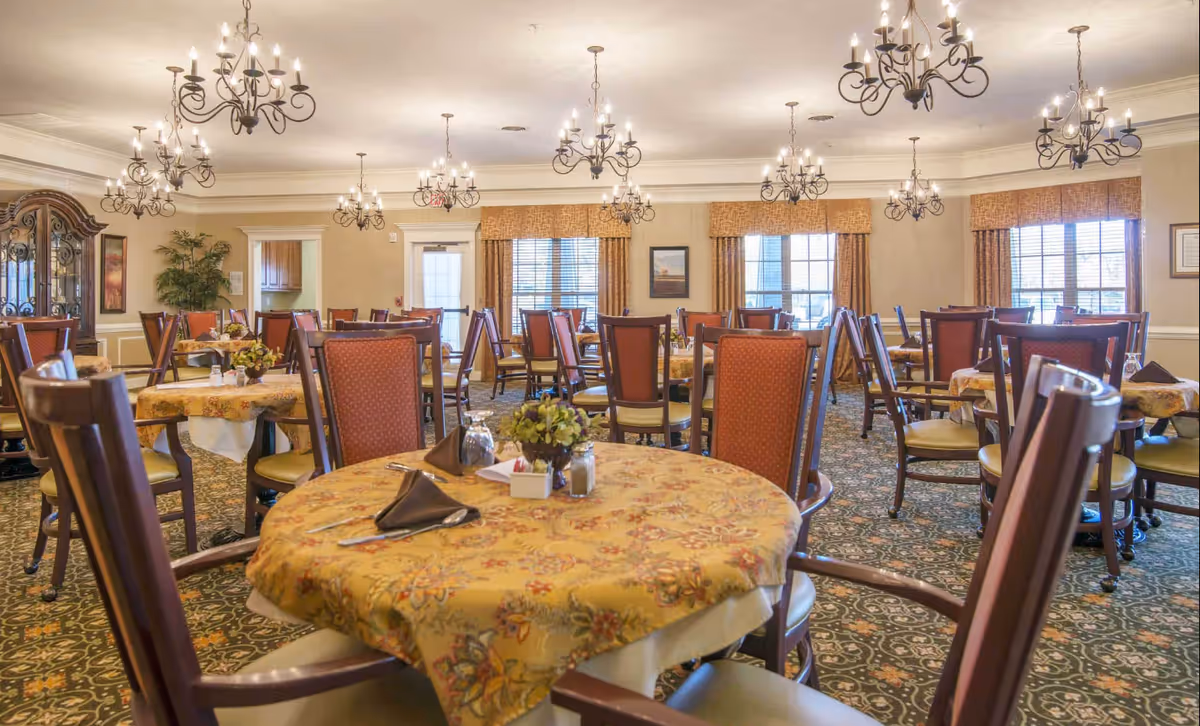 A spacious dining room with multiple round tables covered with yellow floral tablecloths. Each table is set with brown napkins, silverware, and small floral centerpieces. The room features elegant chandeliers hanging from the ceiling, large windows with brown curtains, and a patterned carpet. Wooden chairs with red cushioned backs and light-colored seats surround the tables.