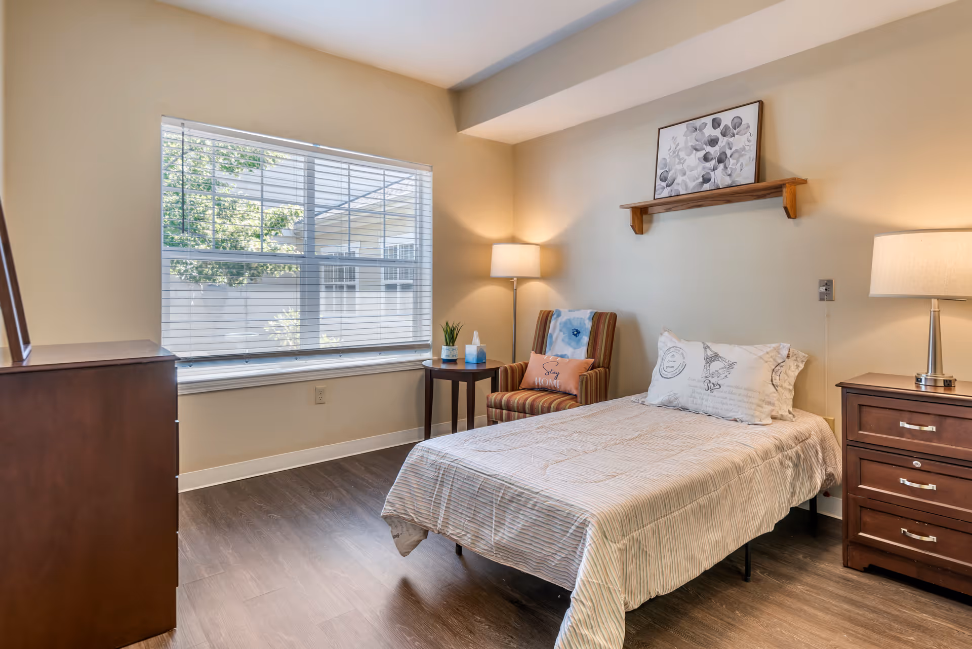 A cozy bedroom with a single bed covered in a light-colored striped bedspread and a decorative pillow. Next to the bed is a wooden nightstand with a lamp. There is a striped armchair with a pillow and a small round table holding a plant and a box of tissues. A large window with blinds lets in natural light, and a framed artwork is displayed on a wooden shelf above the bed.