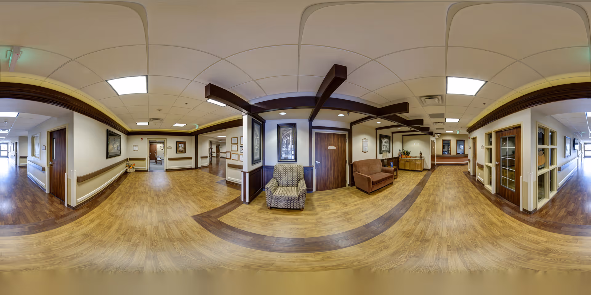 A wide-angle view of a healthcare facility hallway with wood-patterned flooring and white walls accented by dark wood trim. The hallway features several doors, framed artwork, and handrails along the walls. There are two upholstered chairs placed near a wooden cabinet with plants and decorations. The ceiling has recessed lighting and decorative wooden beams. The space is clean, well-lit, and designed for accessibility.