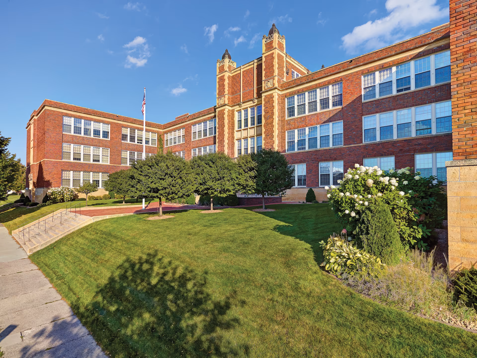 Exterior view of a large brick building with multiple windows, manicured green lawn, trees, shrubs, and a flagpole with an American flag under a clear blue sky.
