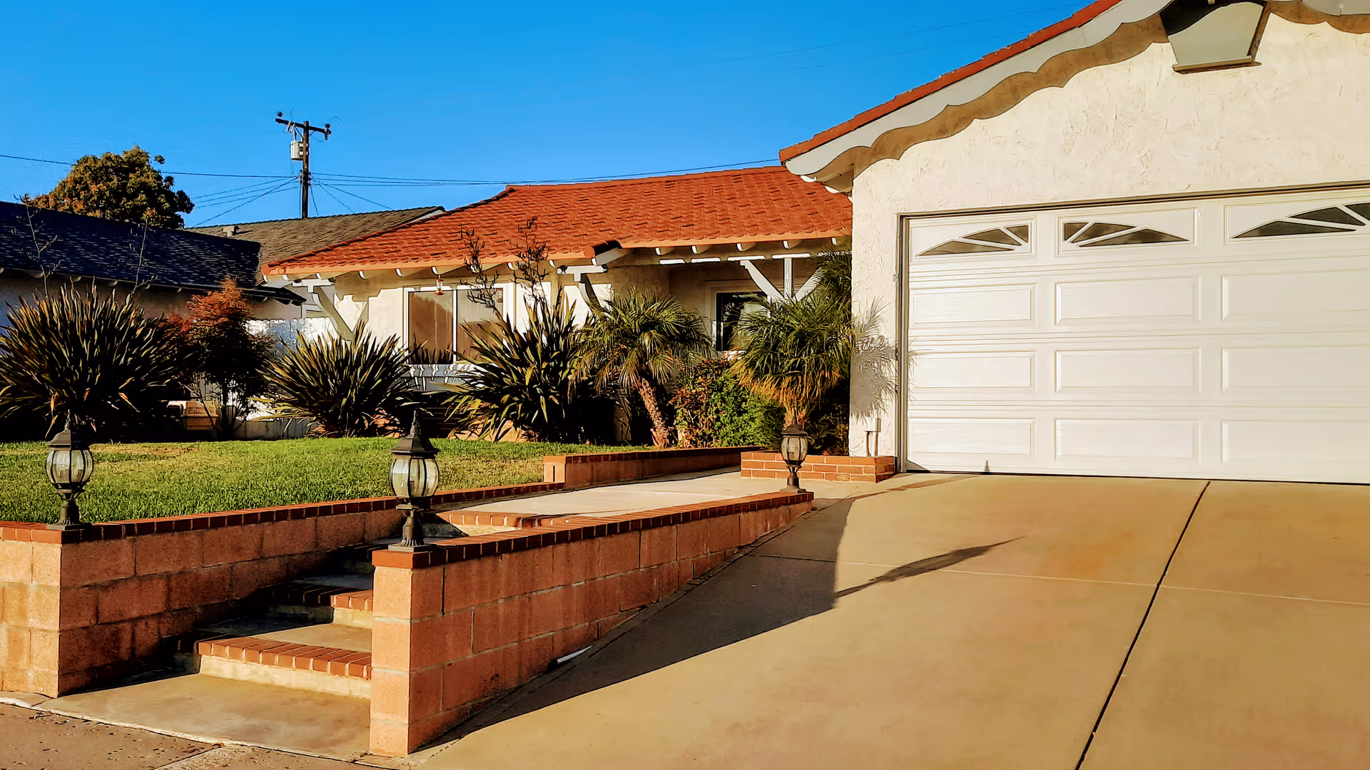 Exterior view of a single-story house with a red-tiled roof, a white garage door, a concrete driveway, and a small front yard with green grass and various shrubs. There are brick steps and a low brick wall with decorative lanterns leading up to the front entrance.