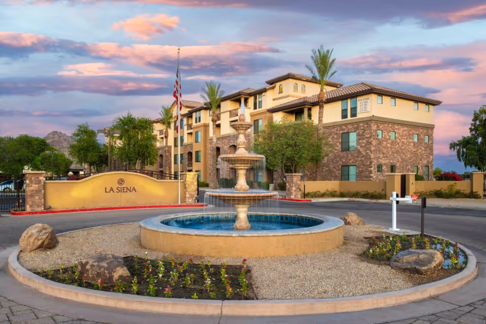 Front entrance of the La Siena building with a central round fountain, landscaped circular drive, and flagpoles under a colorful sky.
