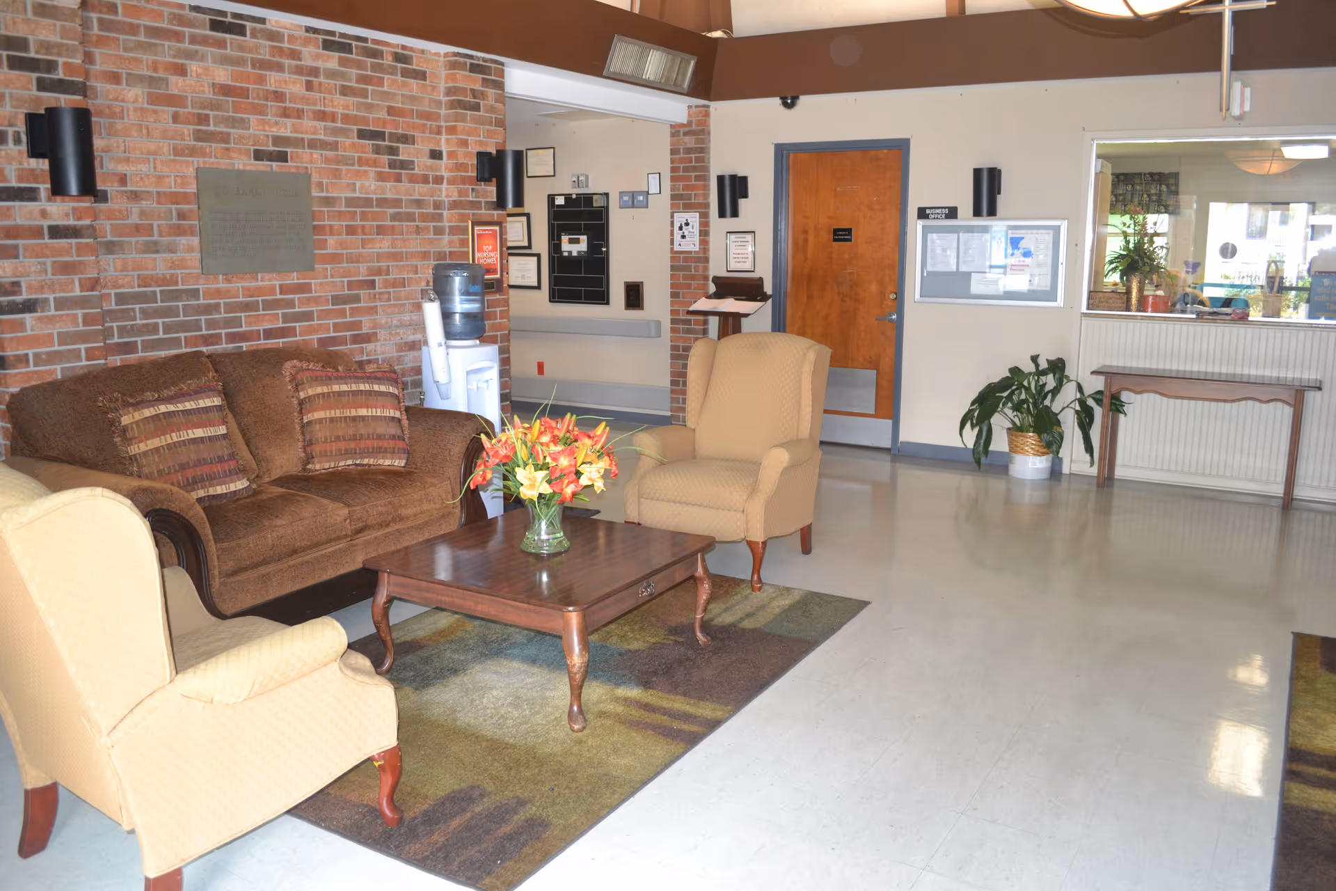A cozy sitting area inside a nursing home with a brown couch, two beige armchairs, a wooden coffee table with a vase of flowers, a brick wall, a water cooler, a wooden door, and a bulletin board on the wall.