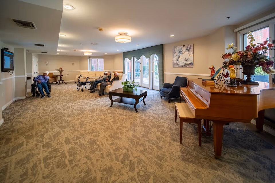 A spacious senior living facility common area with beige patterned carpet and cream-colored walls. There is a wooden grand piano with decorative items and flowers on top to the right. In the middle, a wooden coffee table with a green plant sits in front of a beige sectional sofa where three elderly people are seated. A walker is nearby. To the left, a man in a wheelchair watches a wall-mounted TV. Large windows and glass doors let in natural light, and a floral painting hangs on the wall.