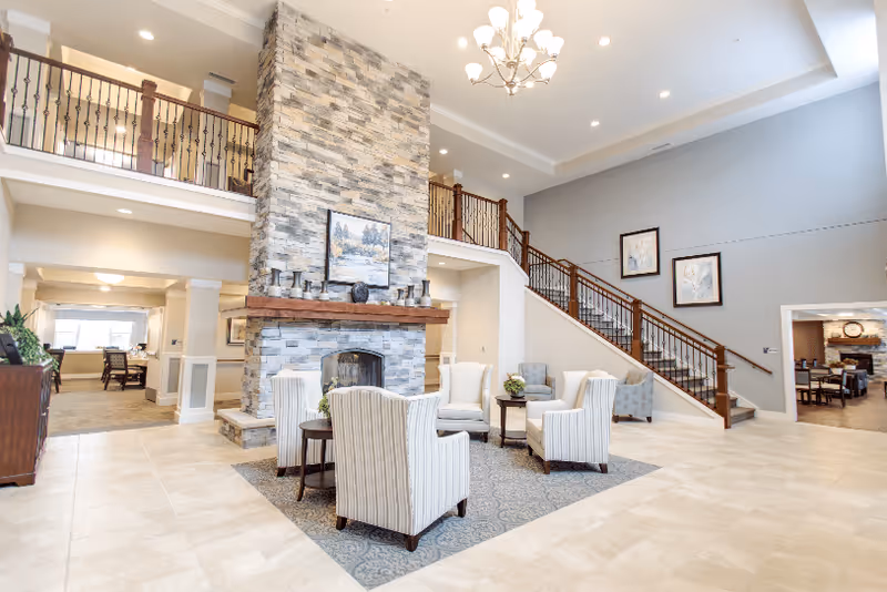 A spacious and well-lit senior living facility common area featuring a large stone fireplace with a wooden mantle, surrounded by comfortable armchairs arranged on a patterned rug. The room has high ceilings with recessed lighting and a chandelier, a staircase with wooden railings leading to an upper level, and framed artwork on the walls. Adjacent rooms and hallways are visible in the background.