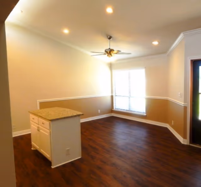 An empty interior room with wood flooring, beige and white walls, a ceiling fan with lights, recessed ceiling lights, a small kitchen island with granite countertop, and a large window with blinds letting in natural light.