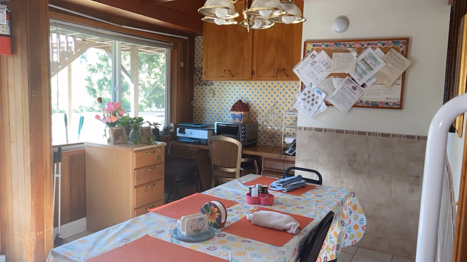 A dining area with a table covered in a colorful tablecloth and four orange placemats. On the table are folded towels, a napkin holder, and condiments. Behind the table is a small desk with a chair, a printer, a toaster oven, and a decorative container. A bulletin board with various papers is mounted on the wall above the desk. A window with a view of greenery is on the left side, and a light fixture hangs from the ceiling.