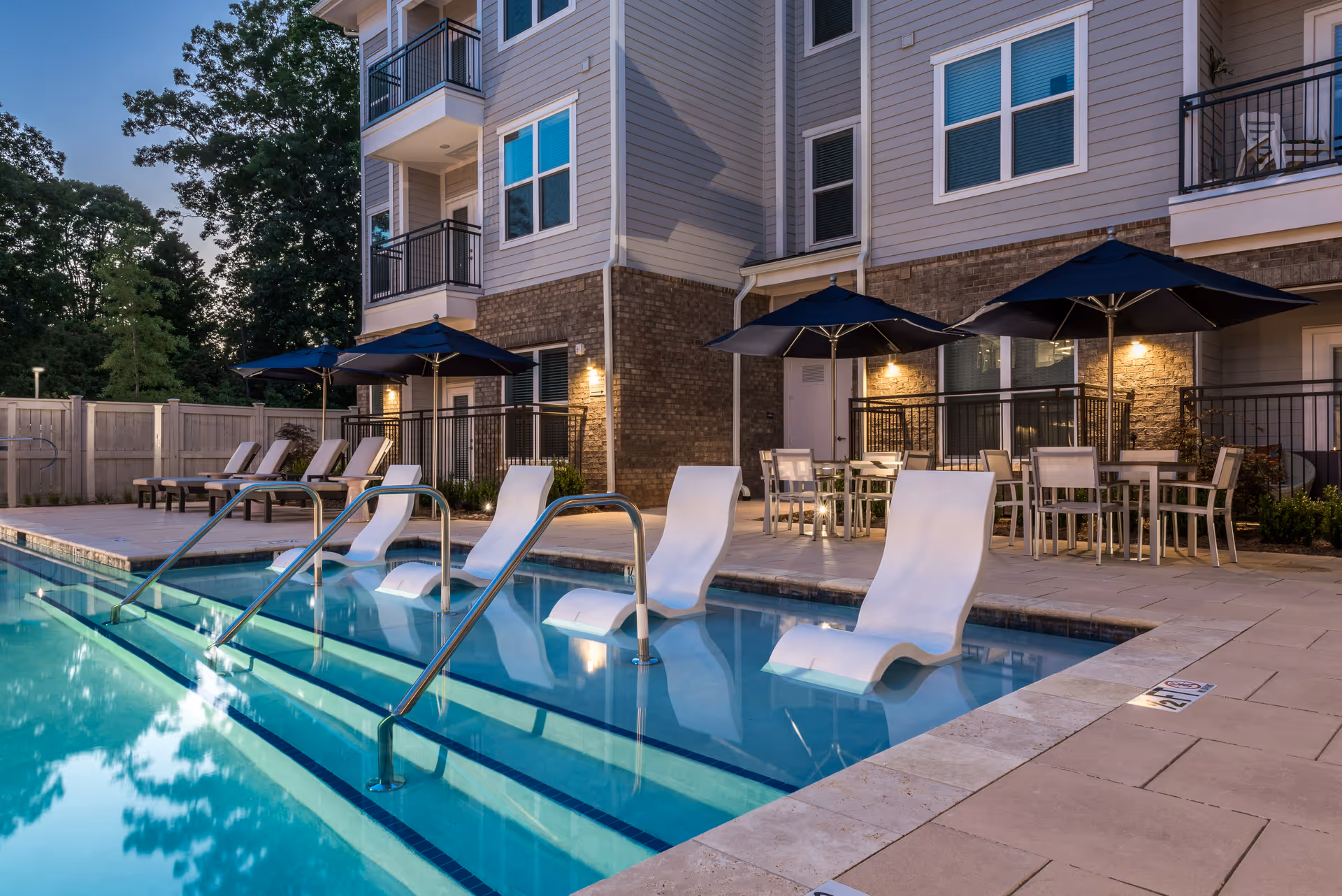 Outdoor swimming pool area at dusk with several white lounge chairs partially submerged in the water, metal handrails, and a poolside patio featuring tables with chairs and large dark umbrellas. The pool is adjacent to a multi-story building with balconies and lit wall sconces.