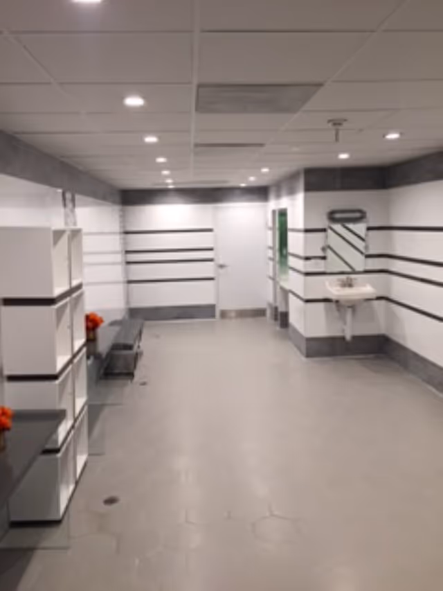 A clean, modern locker room or changing area with white walls featuring horizontal black stripes, a white door at the end, a wall-mounted sink with a mirror, open shelving units, and a bench along the left side. The floor is covered with light gray hexagonal tiles and the ceiling has recessed lighting.