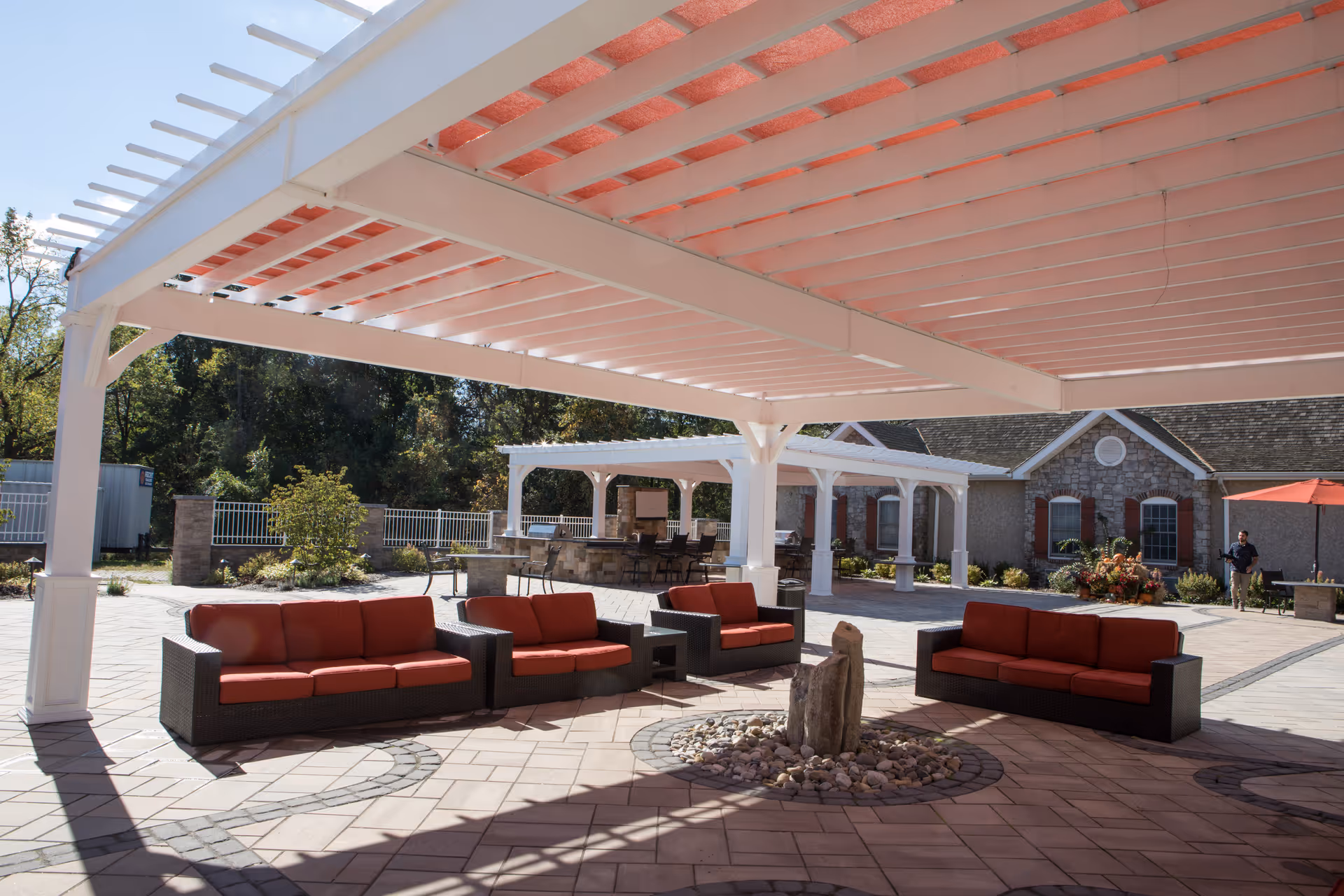 Outdoor patio area with white pergolas providing shade over red cushioned wicker sofas arranged around a small stone fire pit. In the background, there is a stone building with red shutters and a person walking near an umbrella-shaded table. Trees and greenery surround the fenced area.