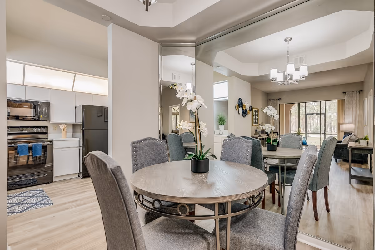 Dining area with a round wooden table and four gray upholstered chairs, decorated with a white orchid centerpiece. The dining area is adjacent to a kitchen with white cabinets, a black refrigerator, and a black stove. A large mirror on the wall reflects the dining area and part of the living room, which has a large window with a view of trees outside.