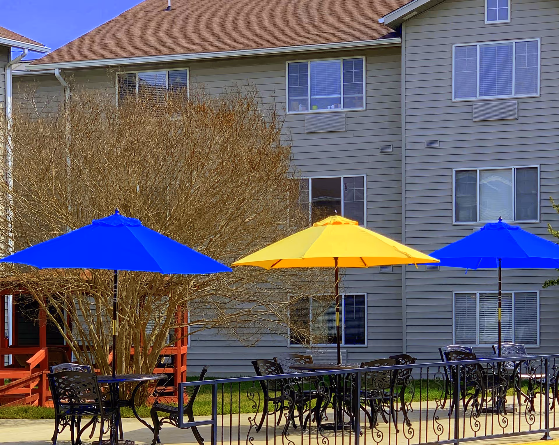 Outdoor patio area with black metal tables and chairs, shaded by two blue umbrellas and one yellow umbrella. In the background, there is a multi-story beige building with several windows and a leafless tree.