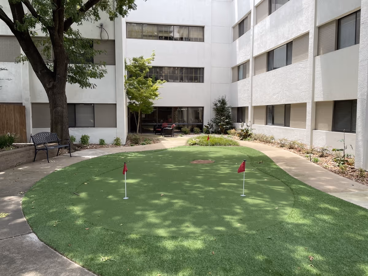 Courtyard with a small artificial putting green with red flags, benches, trees, and seating surrounded by a multi-story white building.