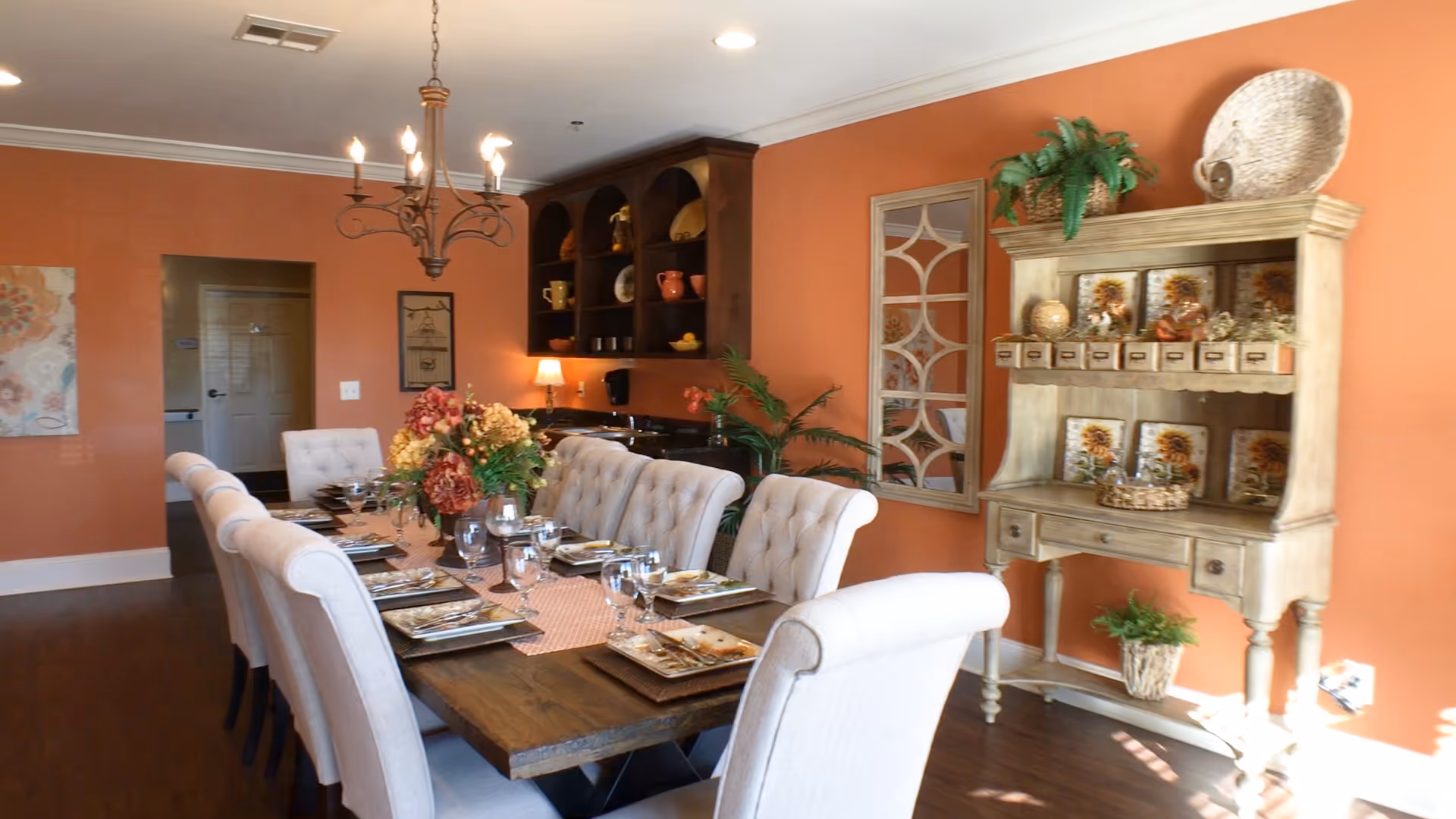 A dining room with a long wooden table set for a meal with plates, glasses, and silverware. The table is surrounded by cushioned beige chairs. The walls are painted orange, and there is a chandelier hanging above the table. A wooden cabinet with decorative items and plants is against the wall, along with a decorative mirror. The floor is dark wood.
